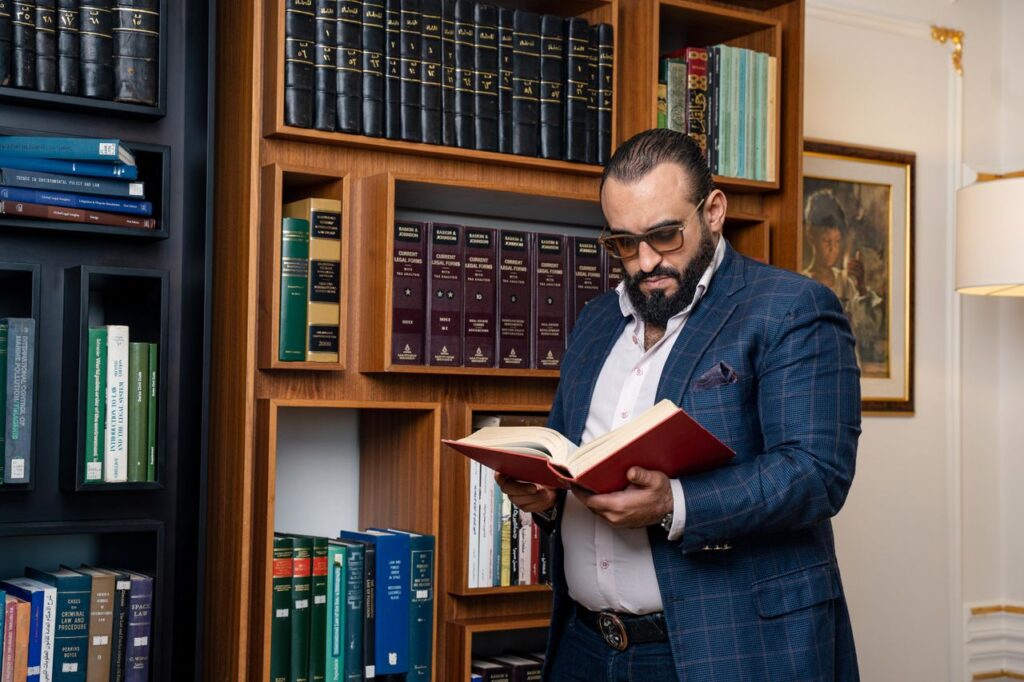 Man with beard and glasses reading a book in a sophisticated library setting.